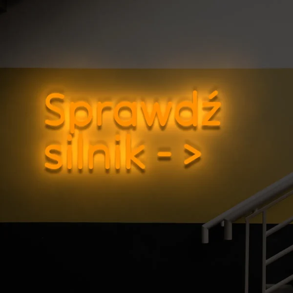 Pomarańczowy neonowy napis „Sprawdź silnik →” na ścianie